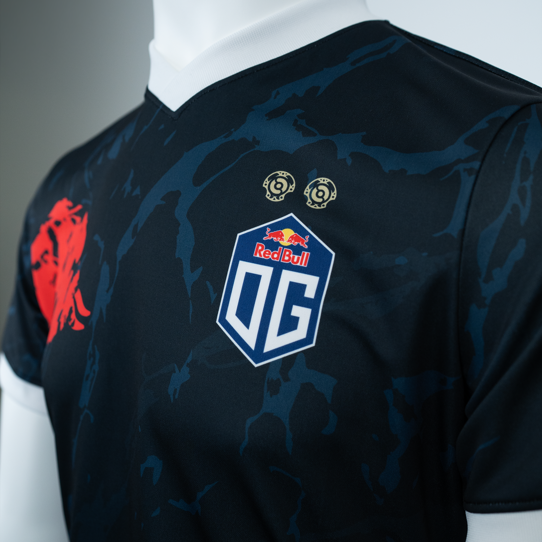 Close-up view of an OG jersey with the OG logo on the left side and a bull logo on the right side, featuring white highlights around the neck and sleeves. The jersey also includes two golden DOTA 2 Aegis symbols above the OG logo.