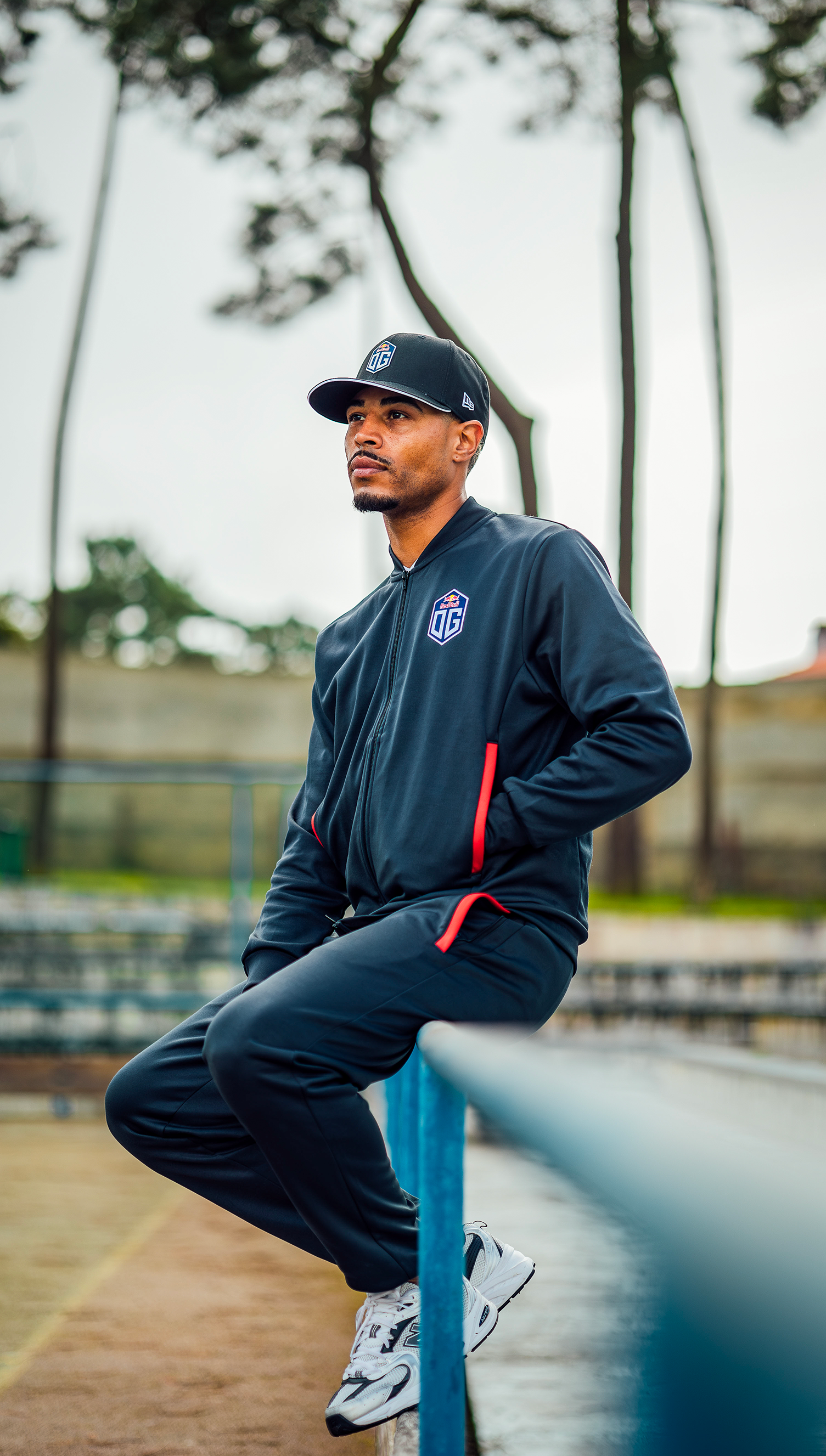 A male model outdoors wearing Esports Team OG apparel: dark grey cap, jersey jacket, and pants, all prominently displaying the OG team logo.