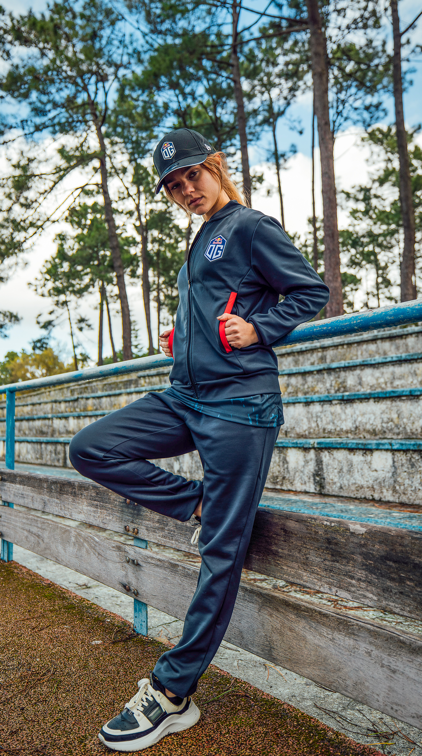 A female model outdoors wearing Esports Team OG apparel: dark grey cap, jersey jacket, and pants, all prominently displaying the OG team logo.