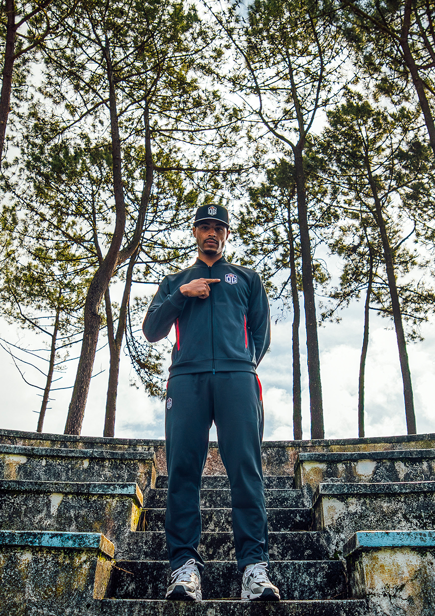 A male model outdoors pointing at the OG logo, dressed in Esports Team OG apparel: dark grey cap, jersey jacket, and pants, all prominently displaying the OG team logo.